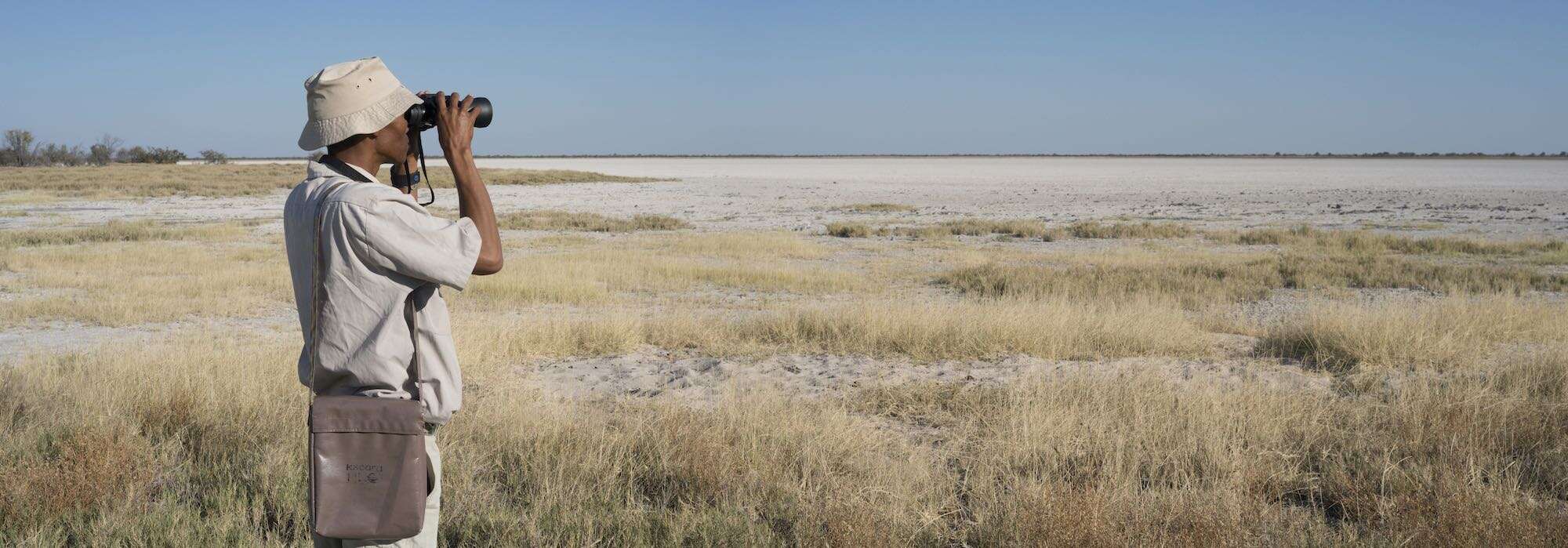 A Namibian ranger looks through binoculars across an open pan.